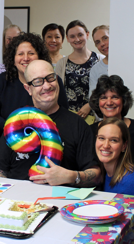 A man holds a rainbow balloon of the number 9. He is surrounded by smiling clinic staff. There is a cake on the table in front of them.