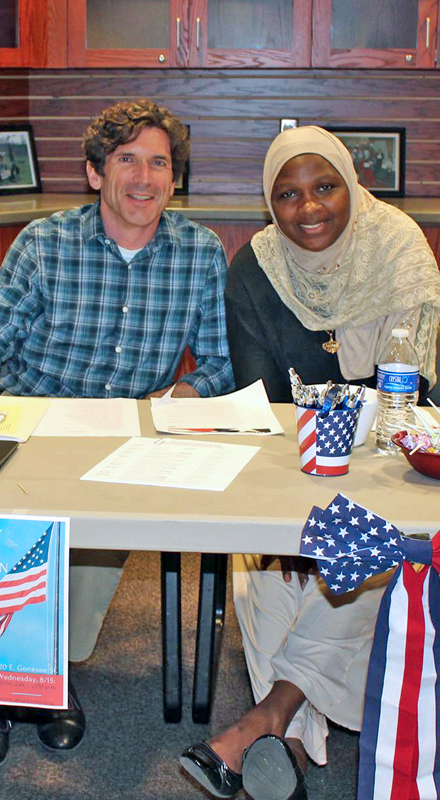 A white man and a Black woman in a headscarf sit at a voter registration table, both are smiling