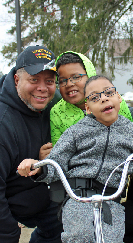 A Black man poses with his two sons, all are smiling, the youngest son is on a tricycle