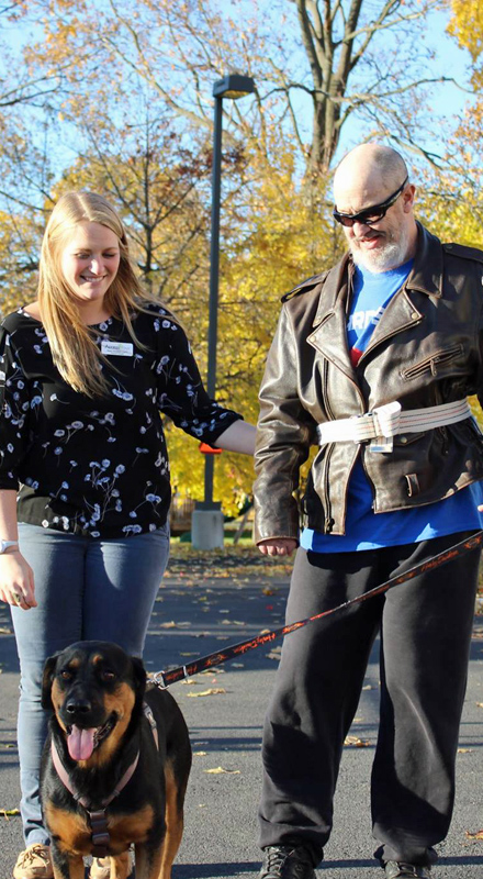 A man in a leather jacket holds a dog by its leash. Next to him an Occupational Therapist provides support. Fall trees can be seen in the background.