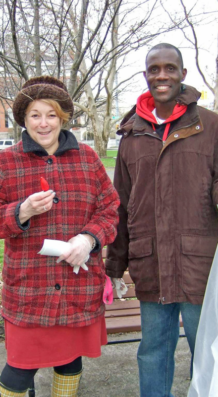 A white woman and a Black man stand outside in winter coats. The woman is holding a Christmas light and the man is holding a bag.