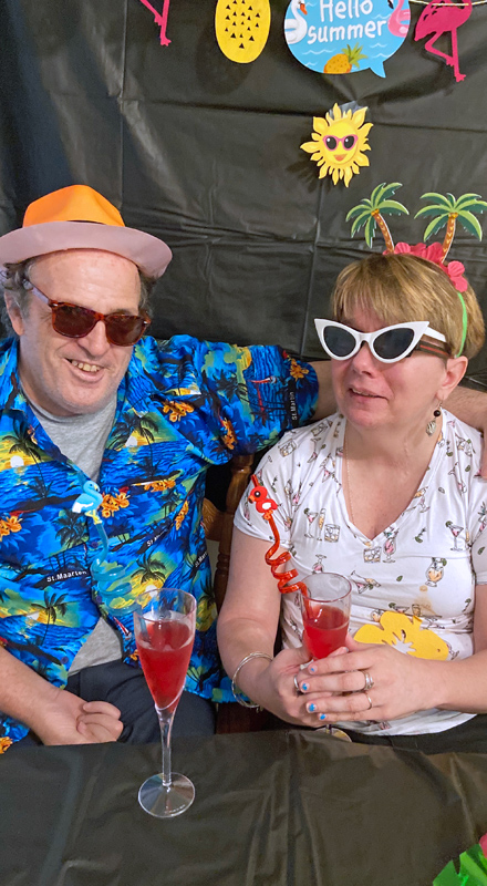 a white man and woman sit together at a table. They are dressed festively in Hawaiian shirts and sunglasses. The woman is holding a red drink with a silly straw