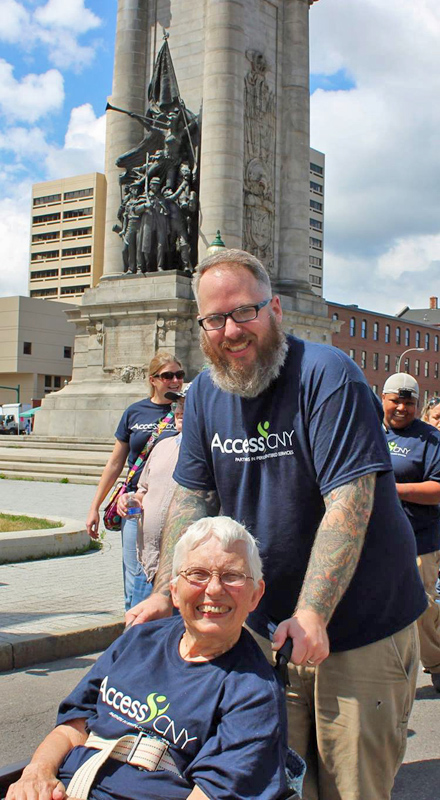 A white man pushes a white woman in a wheelchair. Both are smiling at the camera.