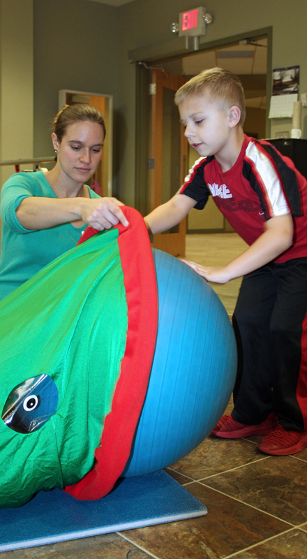 A young white boy pushes an exercise ball through a tube with his physical therapist's help.