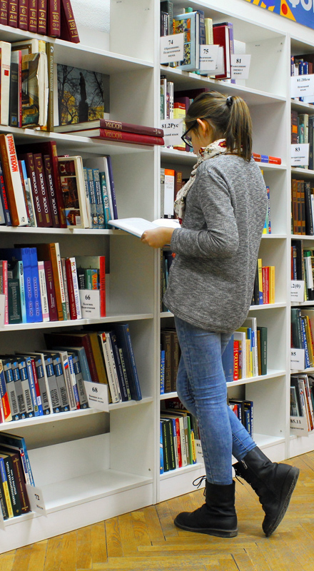 Woman with a ponytail and glasses looks at a book. She stands in front of a white library shelf