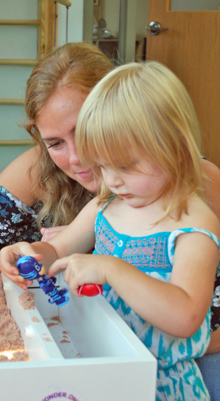 A blonde adult watches as a small blonde girl plays at a sensory table.