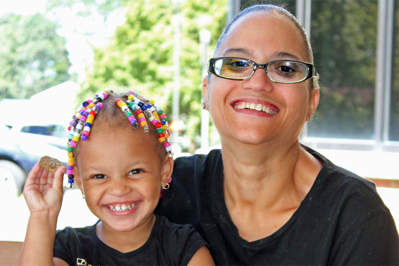 A Black woman with glasses smiles. She is holding her daughter. The daughter has colorful beads in her hair and is holding a rock.