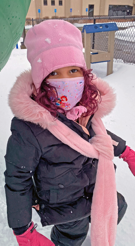 A girl with bright red hair and a pink hat plays in the snow.