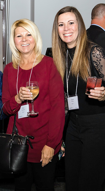 Two white women in formal attire smile at the camera while holding drinks.